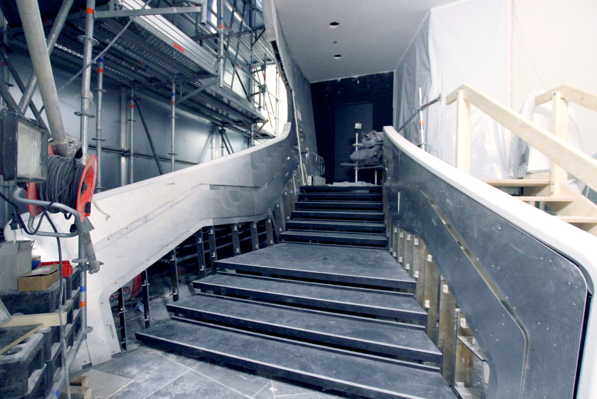 Baustelle einer freigeformten Treppe, die geschwungene Brüstung und der Handlauf stehen roh zwischen Gerüst und Staubschutz, die Stufen sind noch unverkleidet