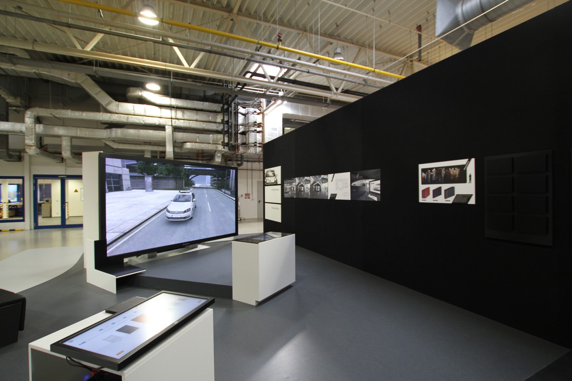 Ausstellungsfläche in einer Werkshalle, schwarze Wand mit Plänen und Mustertafeln, großer Bildschirm mit einem Fahrzeug, weiße Vitrinen unter offen geführter Haustechnik